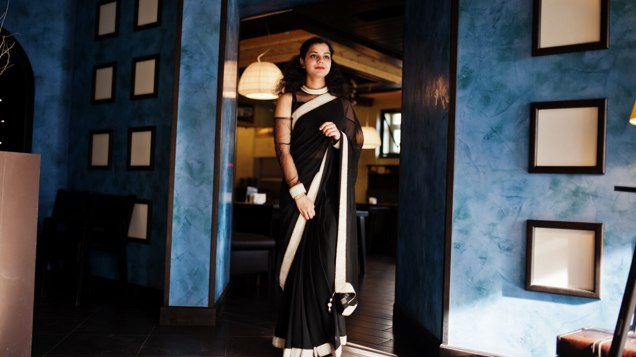 Young indian woman wear at elegant black saree posed on restaurant against wall with frames.